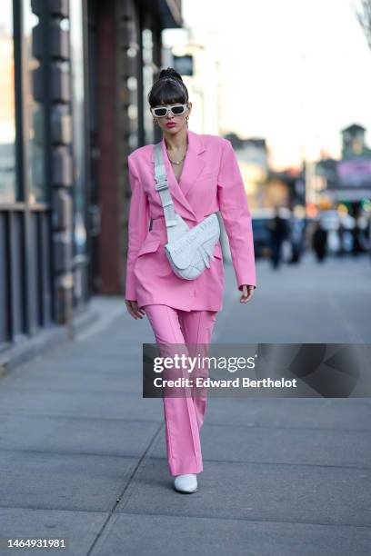 Katya Tolstova wears white sunglasses, gold earrings, a gold large chain necklace, a pink cut-out waist long blazer jacket, matching pale pink wide...