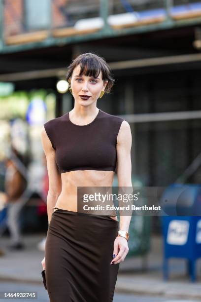 Mary Leest wears gold earrings, a dark brown sleeveless / cropped tank-top, a matching dark brow cut-out waist / gold pearls / long tube skirt,...