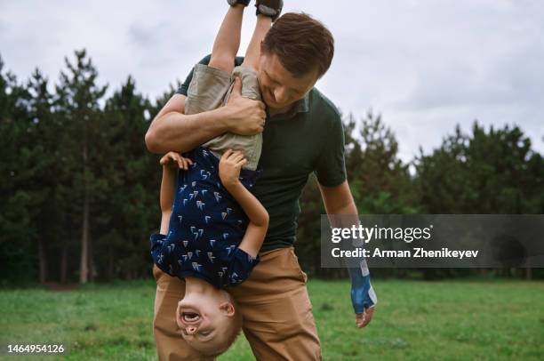 carefree modern father with broken and injured arm wearing orthopedic cast and holding his son upside-down in a natural park - gebrochener arm stock-fotos und bilder