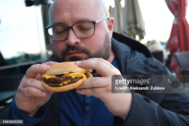 a young man is holding a piece of hamburger in his hands. a bearded guy or man eats fast food. a hungry fat guy is eating an appetizing burger. the concept of junk food, diet, overeating, gluttony, dependence on food. fast food restaurant, snack. - georgian man stock pictures, royalty-free photos & images