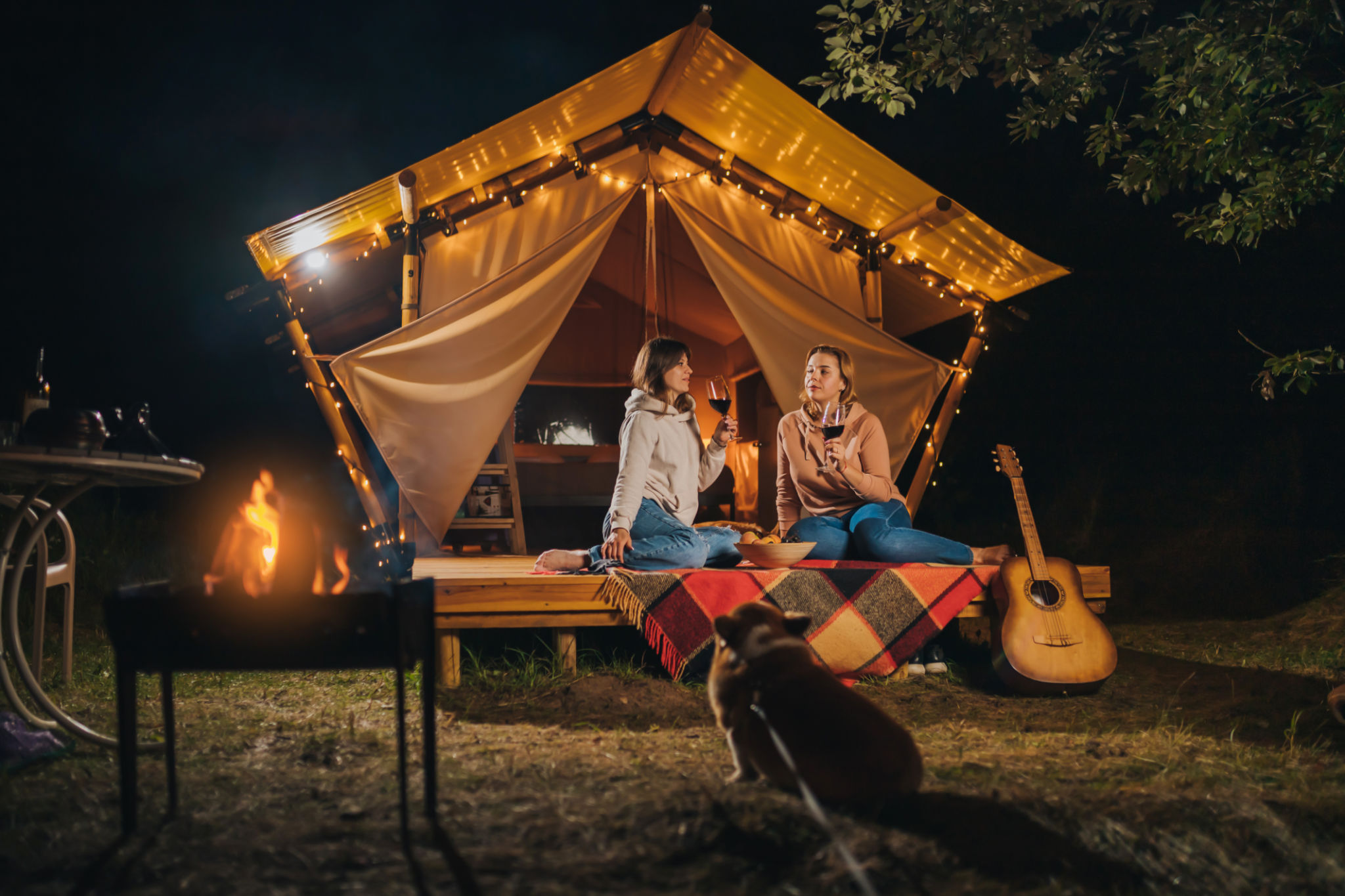 Two Smiling female friends drinking wine and eating fruits sitting in cozy glamping tent in autumn evening bonfire. Luxury camping tent for outdoor holiday and vacation. Lifestyle concept Two Smiling female friends drinking wine and eating fruits sitting in cozy glamping tent in autumn evening bonfire. Luxury camping tent for outdoor holiday and vacation. Lifestyle concept
