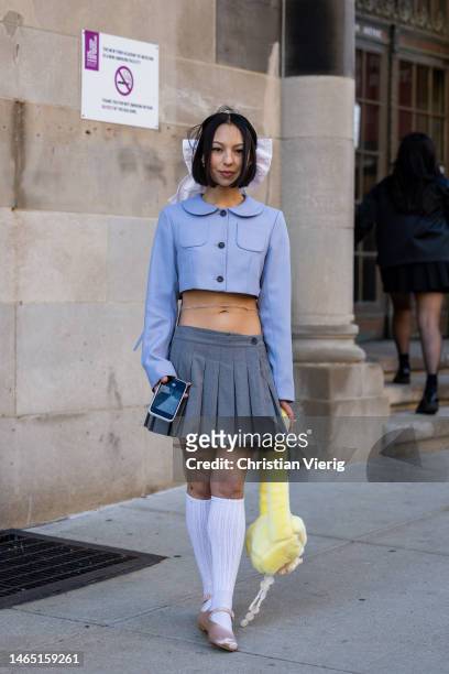 Guest wears cropped jacket, grey pleated skirt, white knee high socks, yellow fleece teddy jacket, bow tie in hair outside Sandy Liang during New...
