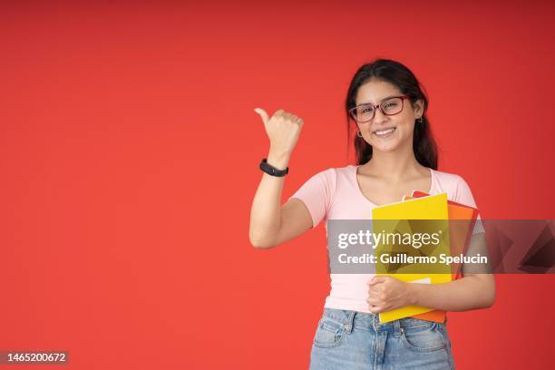 university woman with eyeglasses smiling, posing looking at the camera, with her thumb pointing to the side and the other holding her colored notebooks - happy woman thumbs studio ストックフォトと画像
