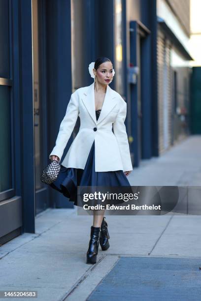 Sai De Silva wears a white flower hair clip, silver earrings, a black lace print pattern shoulder-off corset top / silk ruffled knees dress, a white...