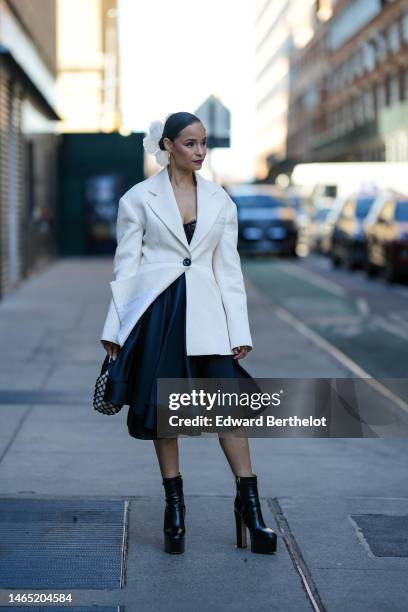 Sai De Silva wears a white flower hair clip, silver earrings, a black lace print pattern shoulder-off corset top / silk ruffled knees dress, a white...