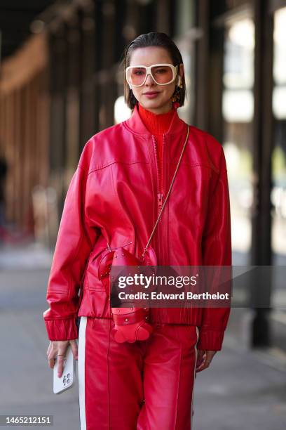 Guest wears white latte oversized sunglasses, gold earrings, a red ribbed wool turtleneck pullover, a red shiny leather bomber coat, a red shiny...