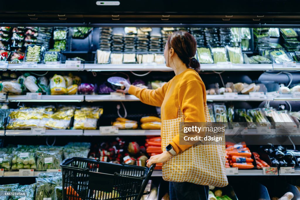 Young Asian woman with shopping cart, carrying a reusable shopping bag, shopping for fresh organic fruits and vegetables in supermarket. Environmentally friendly concept. Zero waste and plastic free. Eco friendly shopping. Sustainable living lifestyle