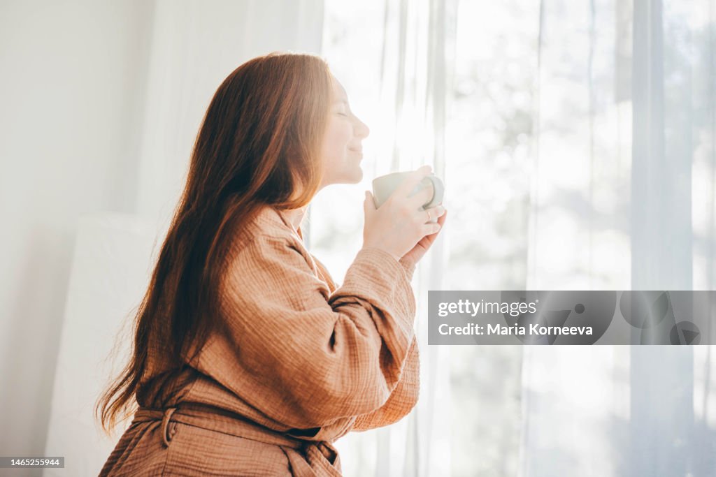 Young woman in bathrobe woken up enjoys the rays of the morning sun and drinking coffee.