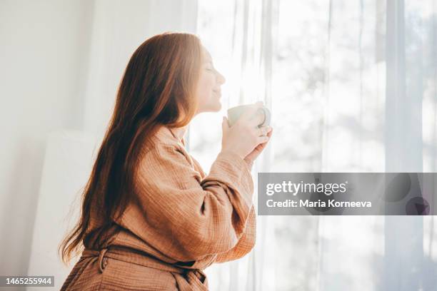 young woman in bathrobe woken up enjoys the rays of the morning sun and drinking coffee. - wake up happy stock pictures, royalty-free photos & images