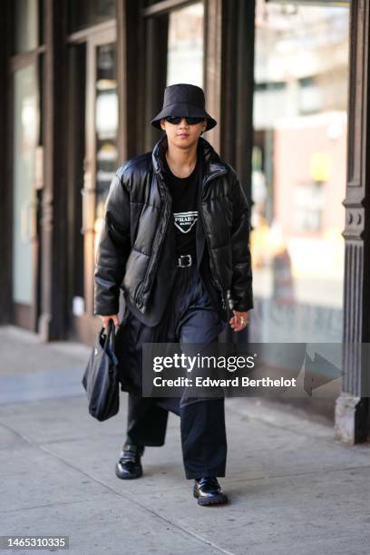 Guest wears a black nylon bob, black sunglasses, gold earrings, a gold chain necklace, a black t-shirt with white logo from Prada, a black shiny...