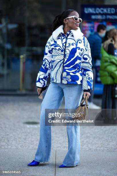 Guest wears white latte sunglasses, a white and royal blue print pattern with white sheep collar zipper coat, blue faded denim wide legs pants, a...