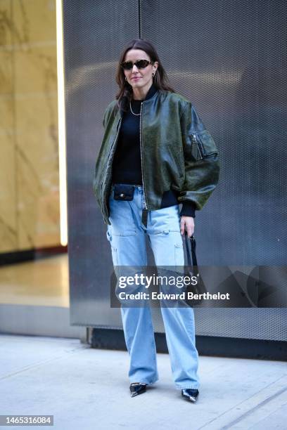 Guest wears brown sunglasses, silver earrings, a black wool pullover, a silver necklace, a dark green shiny leather oversized bomber coat, a black...
