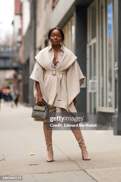 Coco Bassey wears silver earrings, a beige with white print pattern logo silk / short sleeves / knees dress, a beige wool belted oversized cloak, a...