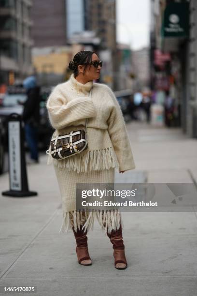 Guest wears brown sunglasses from Miu Miu, gold earrings, gold hair clips, a beige wool ribbed / fringed borders / turtleneck oversized pullover, a...