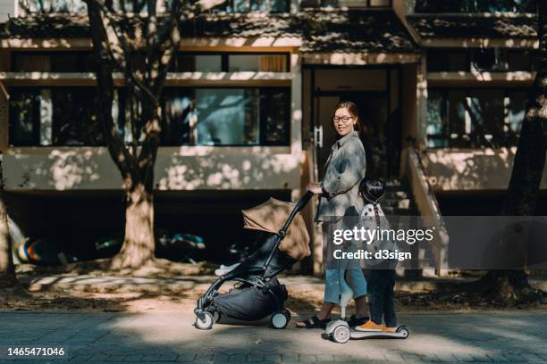 young asian mother pushing a baby stroller, with little daughter by her side riding a scooter. they are having a walk around the neighbourhood on a sunny morning. active family lifestyle. family, child and parenthood concept - step well stockfoto's en -beelden