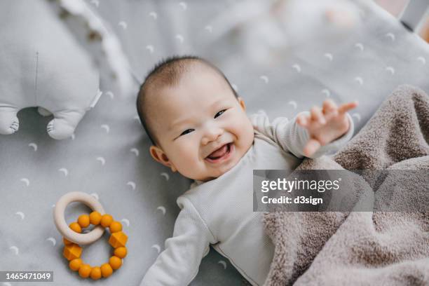 portrait of a lovely asian baby girl smiling sweetly while lying on the crib. she is raising her hand trying to touch the cot mobile. baby's growth and development concept - baby equipment stock-fotos und bilder