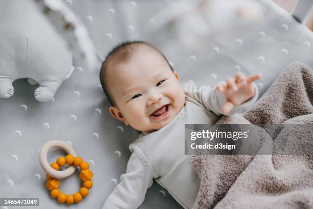 portrait of a lovely asian baby girl smiling sweetly while lying on the crib. she is raising her hand trying to touch the cot mobile. baby's growth and development concept - sólo bebés fotografías e imágenes de stock