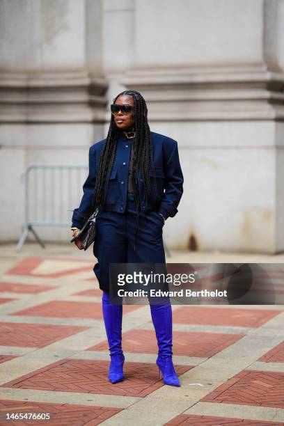 Guest wears black sunglasses, a gold large chain necklace, a black turtleneck pullover, a navy blue buttoned jacket, matching navy blue knees skirt,...
