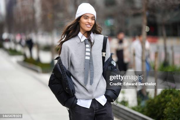 Model wears a white ribbed wool beanie, a white shirt, a gray with large white striped print pattern tie, a pale gray wool pullover, a black puffer...