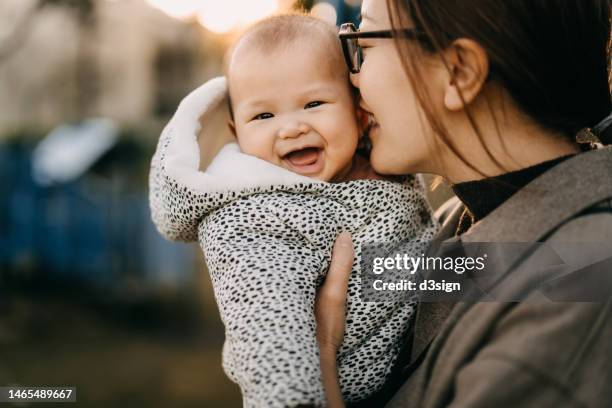 a loving young asian mother carrying her baby girl in arms, kissing on her face, enjoying their time together in the nature. lovely baby girl enjoys it and smiling at camera. love, care and tenderness. family, child and parenthood concept - chinese mothers day foto e immagini stock