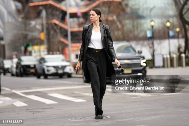Guest wears gold earrings, gold necklaces, a white tank-top, a black shiny leather long coat, a black shiny leather belt, black suit pants, black...