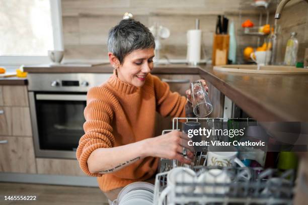 reife frau mit tattoos legt geschirr in die spülmaschine - geschirr spülen stock-fotos und bilder