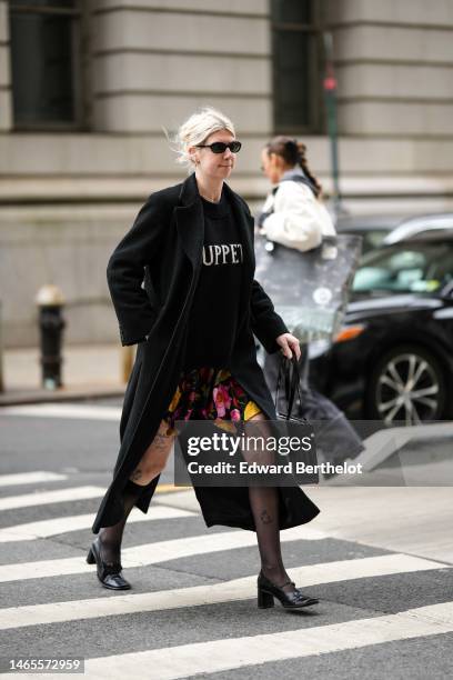 Guest wears black sunglasses, silver earrings, a gold necklace, a black wool with white embroidered inscriptions pattern pullover, a black long wool...