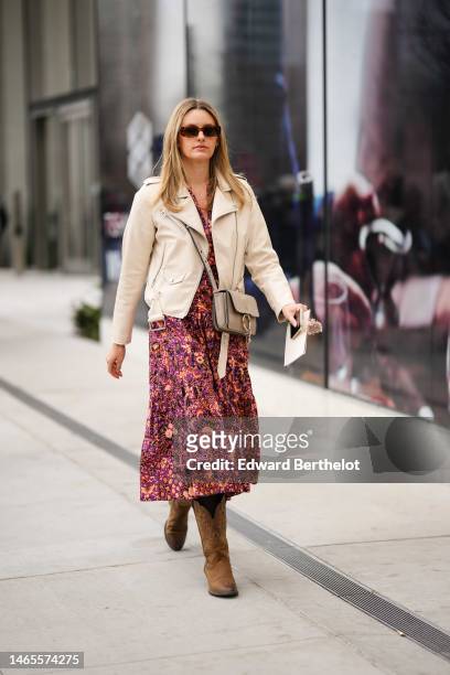 Guest wears brown sunglasses, a purple with black and pale orange flower print pattern V-neck / midi dress, a beige shiny leather zipper jacket, a...