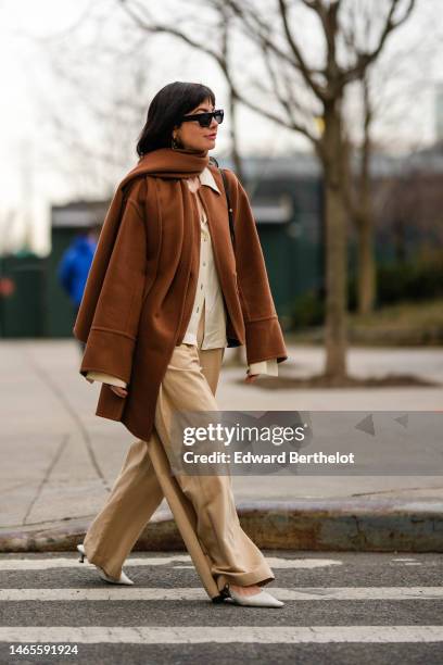 Guest wears black sunglasses, gold large chain pendant earrings, a white shirt, a camel wool oversized jacket, a camel wool long scarf, a black shiny...