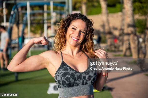 blonde fitness woman with curly hair smiling and pumping biceps outdoors - flexionar los músculos fotografías e imágenes de stock