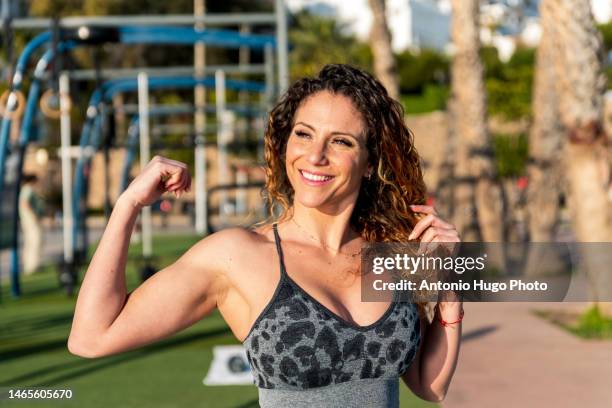 blonde fitness woman with curly hair smiling and pulling out biceps - flexionar los músculos fotografías e imágenes de stock