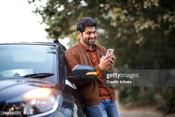 smiling young man texting on mobile phone leaning on her car - leunen stockfoto's en -beelden