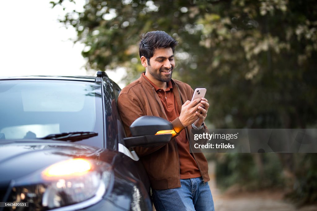Smiling young man texting on mobile phone leaning on her car