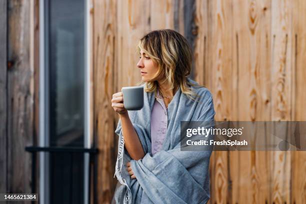 a happy beautiful blonde woman in pajamas contemplating while enjoying her morning coffee - wrapped in a blanket stock pictures, royalty-free photos & images