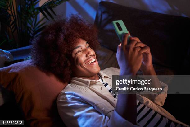 happy young african man lying on the sofa at night, using mobile phone to send online texts. - dating online stock pictures, royalty-free photos & images