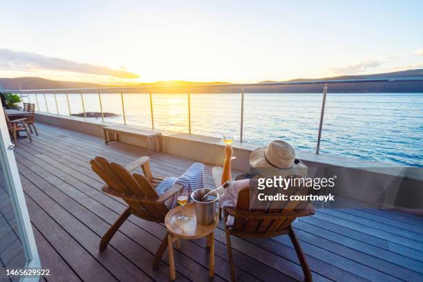 woman relaxing and drinking wine on deck chairs in an over water bungalow. - beach balcony stock pictures, royalty-free photos & images