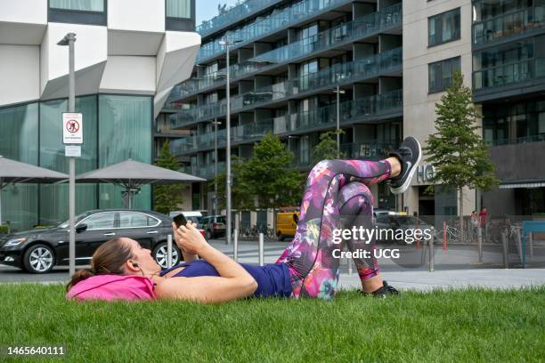 Casual dressed young woman lying, cross-legged, on grass in city center using smartphone. Dublin, Republic of Ireland, Europe, European Union, EU..