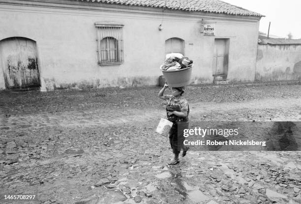 Woman, wearing an tradtional traje, balances a laundry basket on her head as she walks past a pharmacy, south-central Guatemala, July 1982.