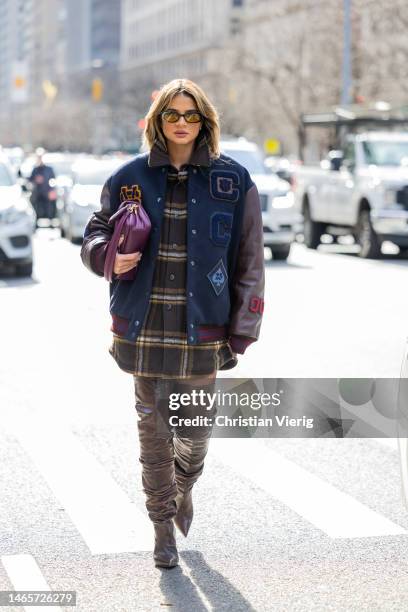 Thassia Naves wears college jacket, checkered button shirt, brown over knees boots, purple bag outside Coach during New York Fashion Week on February...