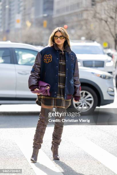 Thassia Naves wears college jacket, checkered button shirt, brown over knees boots, purple bag outside Coach during New York Fashion Week on February...