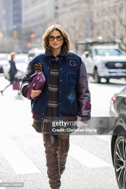Thassia Naves wears college jacket, checkered button shirt, brown over knees boots, purple bag outside Coach during New York Fashion Week on February...