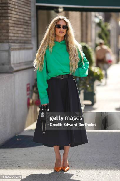 Emili Sindlev wears black sunglasses, a green hoodie sweater, a black shiny leather belt, a black pleated midi skirt, a black shiny leather with...