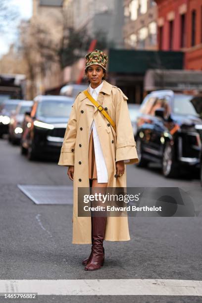 Guest wears a green / brown / beige braided wool hat, gold earrings, a white shirt, a beige long trench coat, a yellow shiny leather crossbody bag...