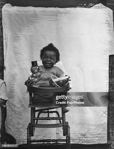 Young girl in a highchair is posed for a photographer with a toy duck, 1930.