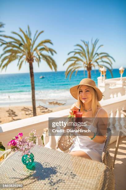 young blonde woman sitting in the beach restaurant on a table enjoying with a smile a red cocktail with the turquoise sea  in the background - blue white summer hat background stock pictures, royalty-free photos & images