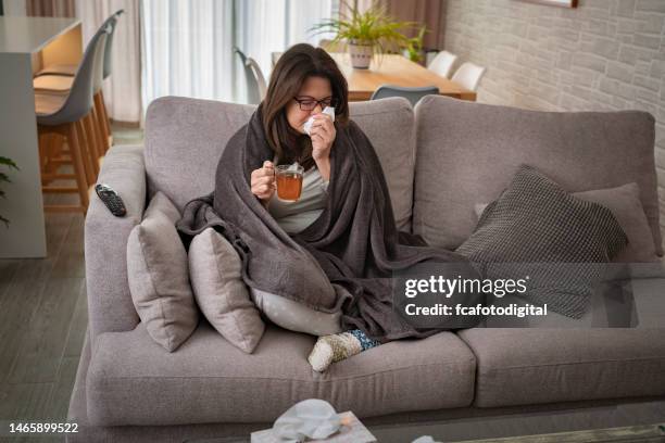 sick woman sneezing in a napkin, sitting on sofa and holding hot tea cup - drinking cold drink stock pictures, royalty-free photos & images