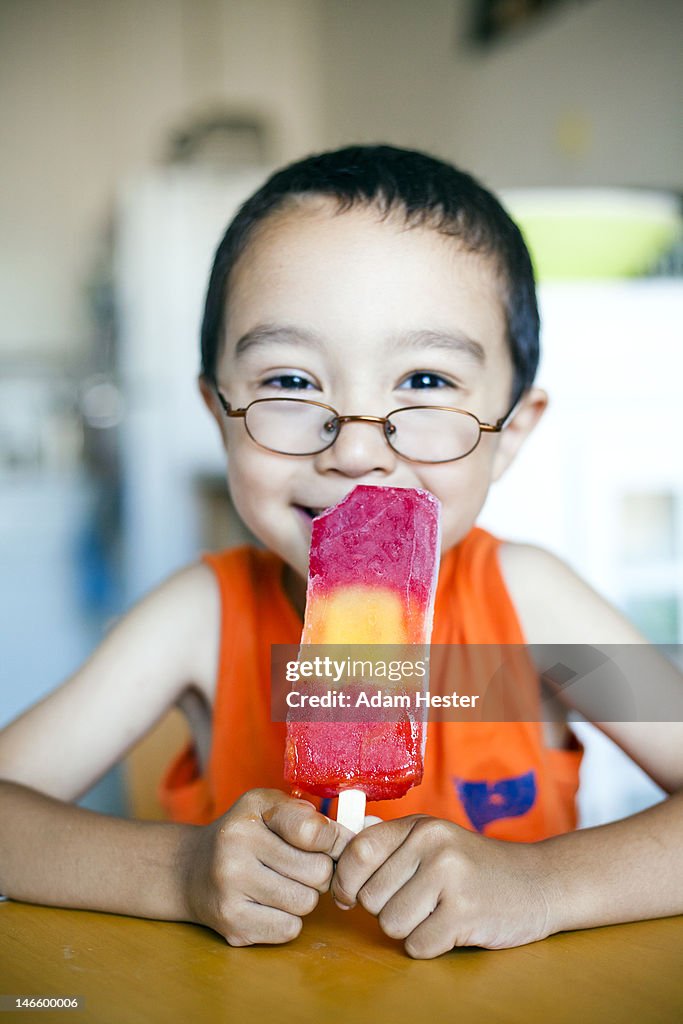 A young boy eating a popsicle inside a kitchen.