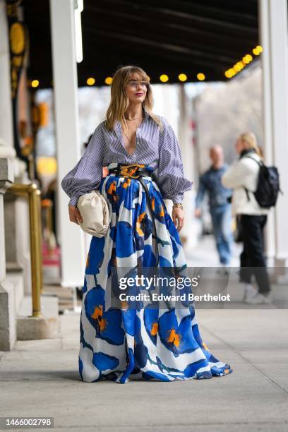 Guest wears glasses, gold chain necklaces, a navy blue and white striped print pattern shirt, a white with royal blue and orange flower print pattern...