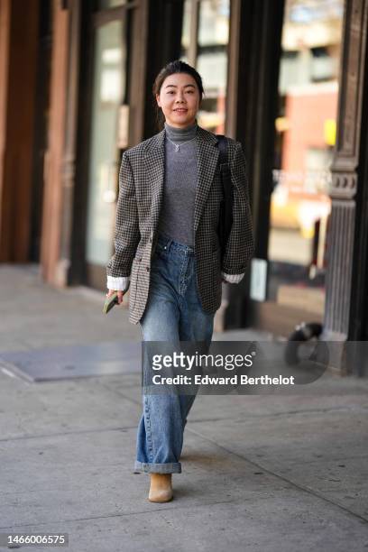 Guest wears a gray turtleneck pullover, a dark brown and black checkered print pattern blazer jacket, a black shiny leather shoulder bag, blue faded...