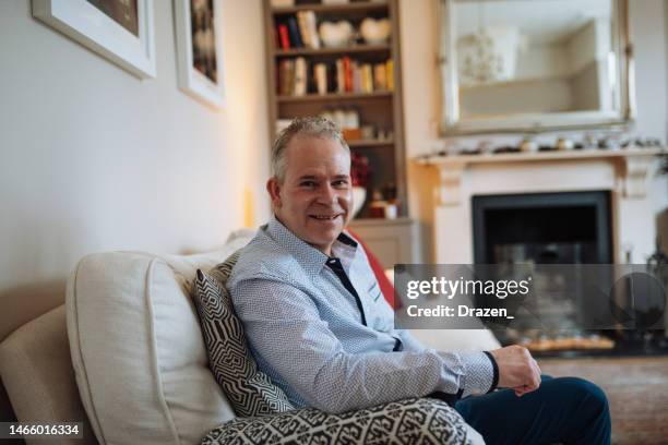 a day in life of smiling senior man sitting on sofa and looking at camera in traditional living room with fireplace - day in the life series stock pictures, royalty-free photos & images